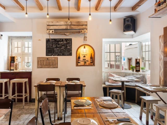 Sala com vigas de madeira, mesas altas e um nicho iluminado com garrafas de vinho